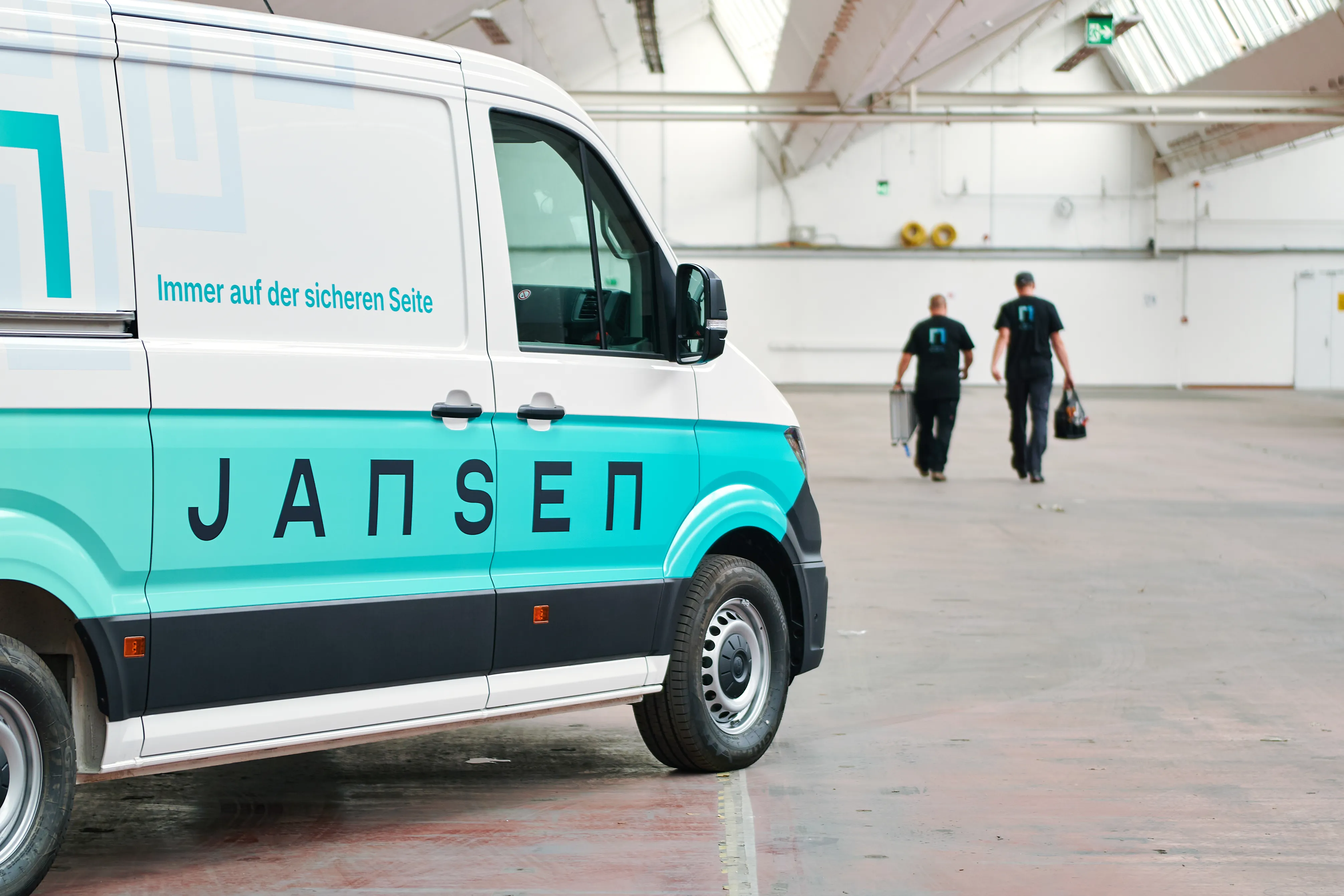 Links steht, in einer Halle, ein Transporter mit der Aufschrift der Jansen Group. Rechts im Hintergrund sind zwei Mitarbeitende von hinten zu sehen, auf deren T-Shirts das Firmenlogo abgebildet ist.