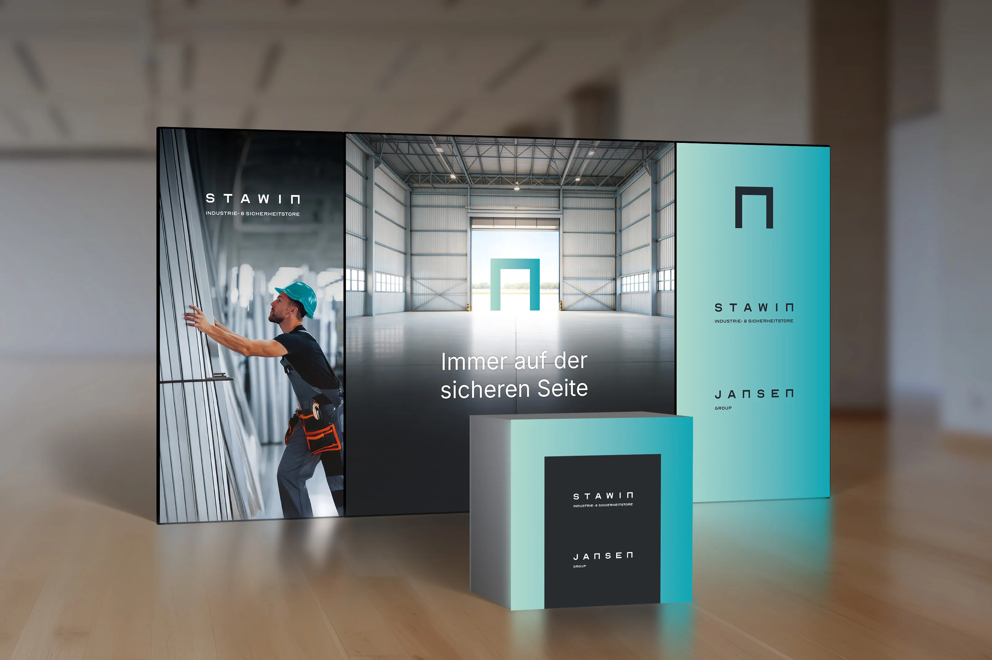 Eine dreiteilige Werbewand. Auf der linken Seite der Wand ist ein Arbeiter in einem blauen Helm zu sehen. Die mittlere Leinwand zeigt eine große Halle mit einem geöffneten Tor und dem Slogan „Immer auf der sicheren Seite“ in weißer Schrift. Auf der rechten Seite sind die Logos von Stawin und Jansen Group auf einem blauen Hintergrund zu sehen. Auf dem Holzboden davor steht ein kleinerer, quadratischer Block, der ebenfalls mit den Logos und dem gleichen blauen Farbverlauf bedruckt ist.