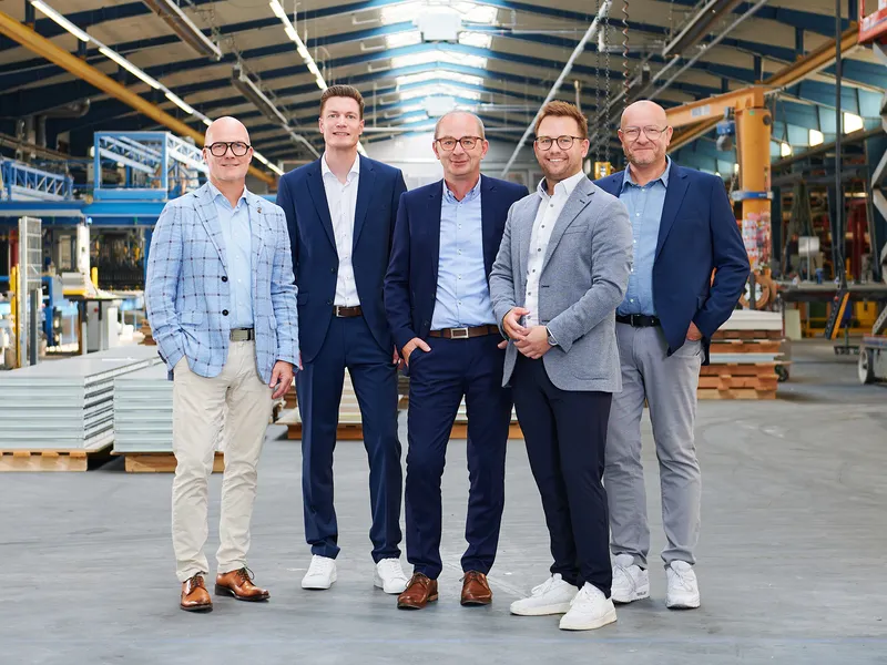 Das Führungsteam, bestehend aus fünf Männern in Anzügen, stehen in einer Produktionshalle und lächeln in die Kamera.